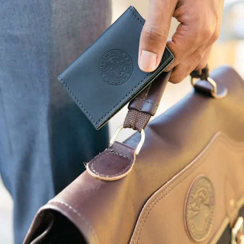 Business Card Holder in premium black leather with embossed logo, held alongside a brown leather and canvas briefcase, reflecting heritage craftsmanship by Duluth Pack.