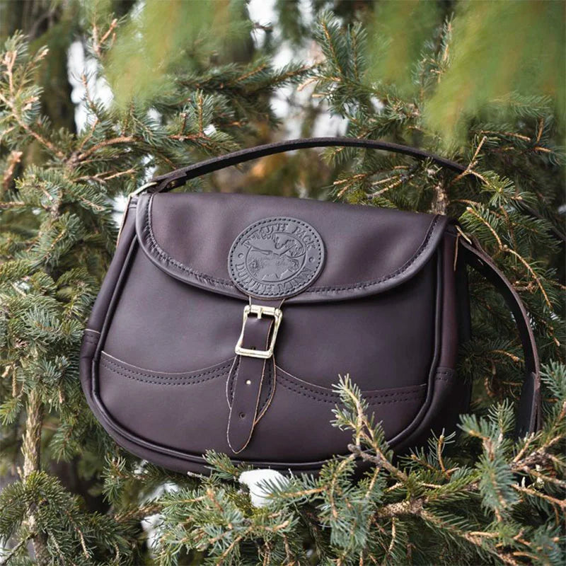 Medium Leather Shell Purse in dark brown premium leather with antique brass buckle, nestled among green pine branches, showcasing durable craftsmanship for outdoor use.