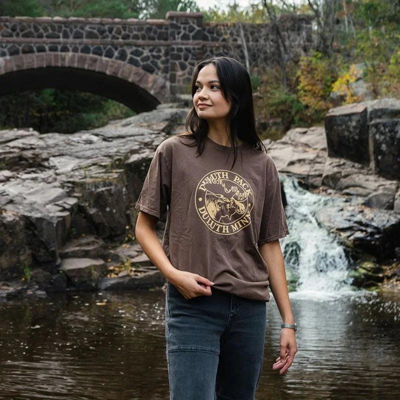 Duluth Pack Short Sleeved Logo T-Shirt in brown cotton, featuring a classic logo, modeled outdoors near a rocky waterfall and stone bridge by LAKESHIRTS, INC.