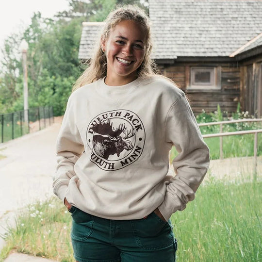 Duluth Pack Logo Crewneck in natural cotton blend featuring iconic graphic, styled casually outdoors by a smiling woman in front of rustic wooden building.