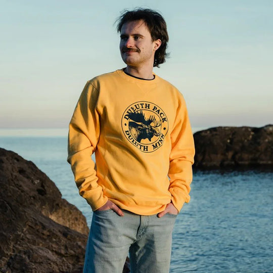 Duluth Pack Logo Crewneck in warm yellow featuring iconic logo graphic, worn casually by a man outdoors near rocky coastline during sunset.