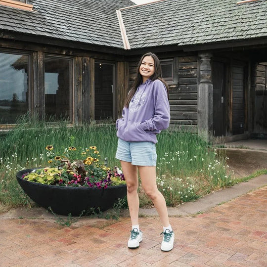 Duluth Pack Logo Sweatshirt in soft purple worn by a smiling woman outdoors near rustic wooden building, showcasing comfortable outdoor apparel by LAKESHIRTS, INC.