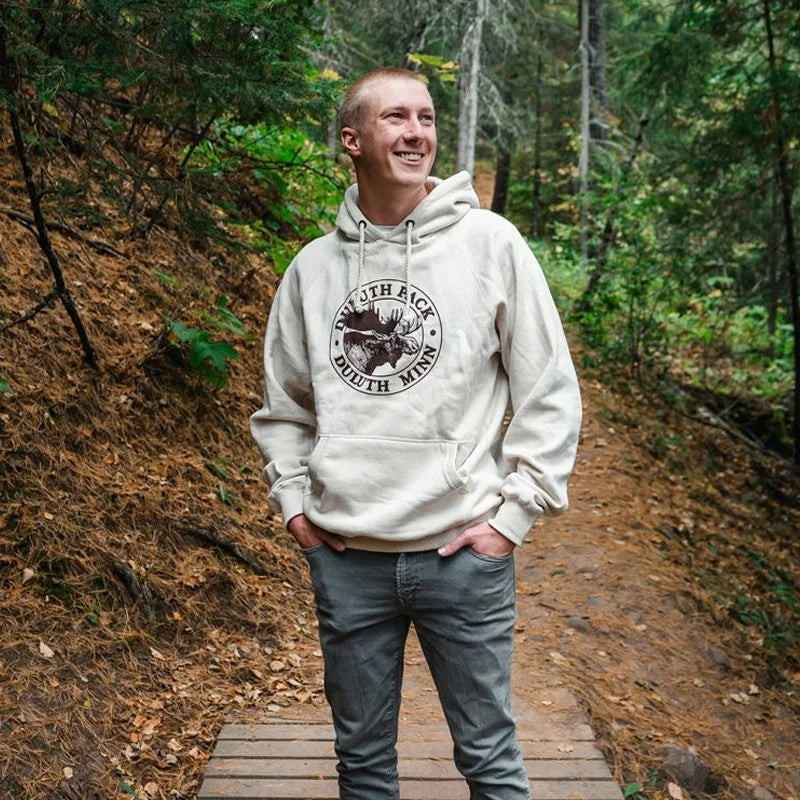 Duluth Pack Logo Sweatshirt in cream with iconic logo, worn by a smiling man outdoors on a woodland trail, showcasing handmade quality by LAKESHIRTS, INC.
