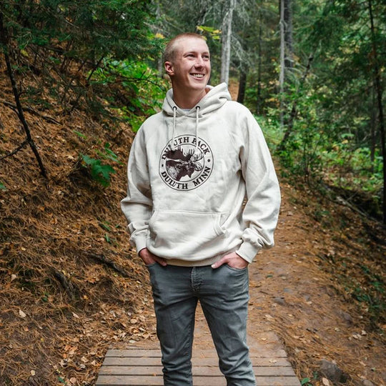 Duluth Pack Logo Sweatshirt in cream with iconic logo, worn by a smiling man outdoors on a woodland trail, showcasing handmade quality by LAKESHIRTS, INC.
