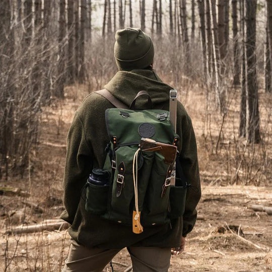 Outlander Pack in green canvas and leather, designed for compact bushcraft use on narrow trails, worn by a hiker in a dense forest setting.