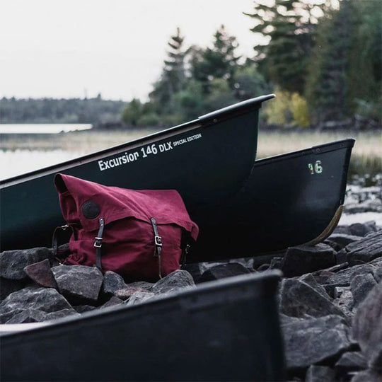 #3 Original Pack - 72 L in maroon canvas with rugged leather straps placed on rocky shoreline beside canoes in an outdoor wilderness setting