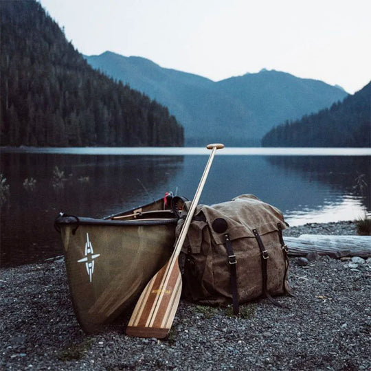 #4 Monarch Pack in burly 15-ounce canvas with leather accents, resting beside a canoe and wooden paddle at a lakeside with forested mountains in the background, showcasing rugged outdoor gear for durable canoe travels.