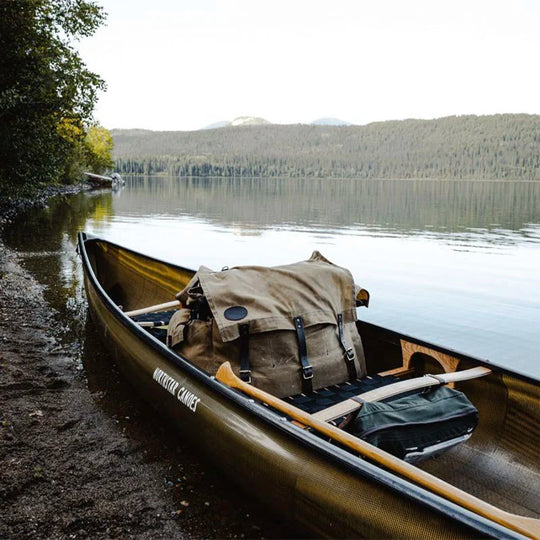 #4 Monarch Pack in burly canvas and leather resting inside a traditional canoe on a calm lake shore, reflecting heritage craftsmanship by Duluth Pack.