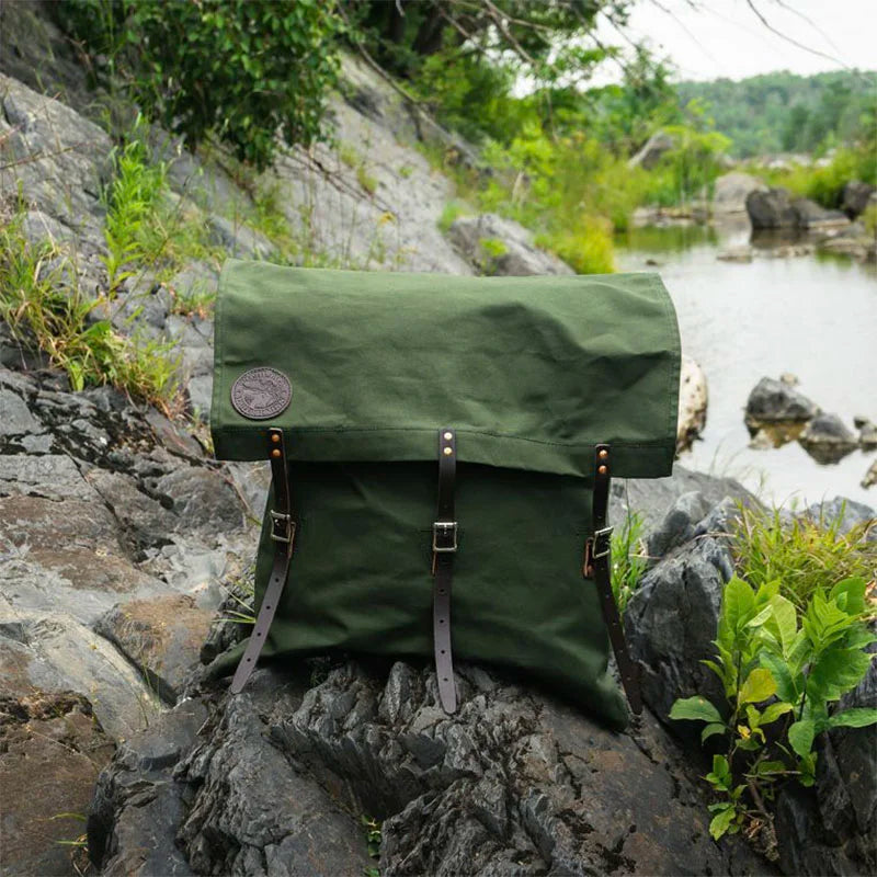 #60 Utility Pack - 42 L in Olive Drab canvas with leather straps, handcrafted by Duluth Pack, resting on rocks beside a peaceful river in a natural outdoor setting.