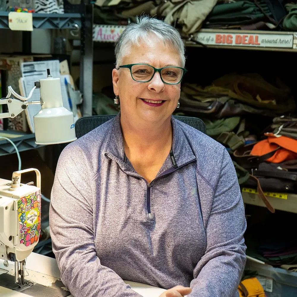 Experienced artisan in a purple pullover sitting at a sewing machine in a workshop filled with canvas and leather materials, highlighting traditional handcrafted gear.