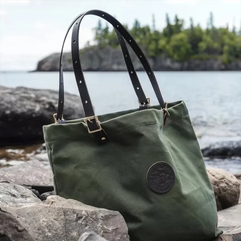 Market Tote in Olive Drab with adjustable leather handles, rugged 15oz canvas construction, photographed outdoors by a lakeside rock setting.