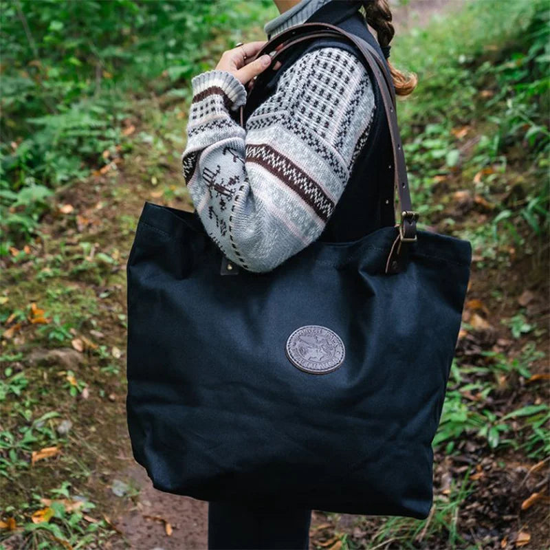Market Tote in black rugged canvas with adjustable leather handles, carried outdoors on a forest trail, showcasing durable handmade craftsmanship by Duluth Pack.