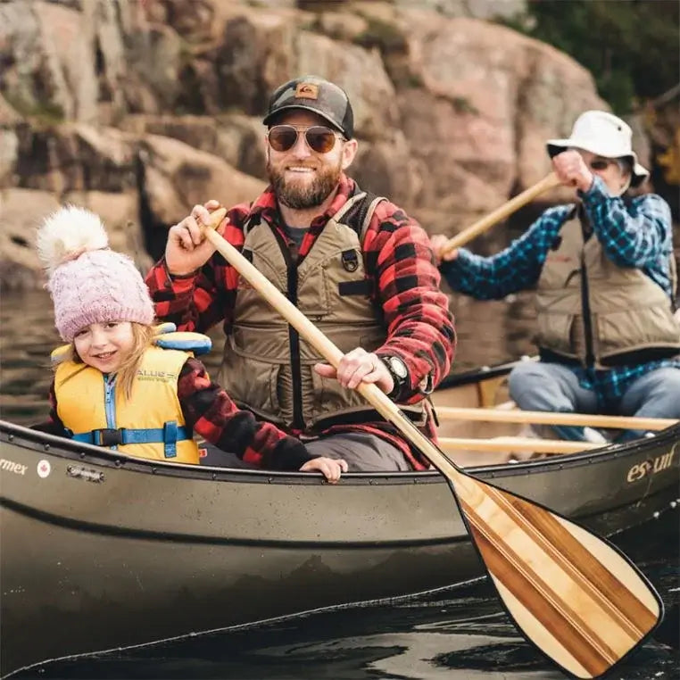 Bending Branches Expedition Plus Paddle Being used by a man in a canoe with another person and a child inside on a lake with a rock background