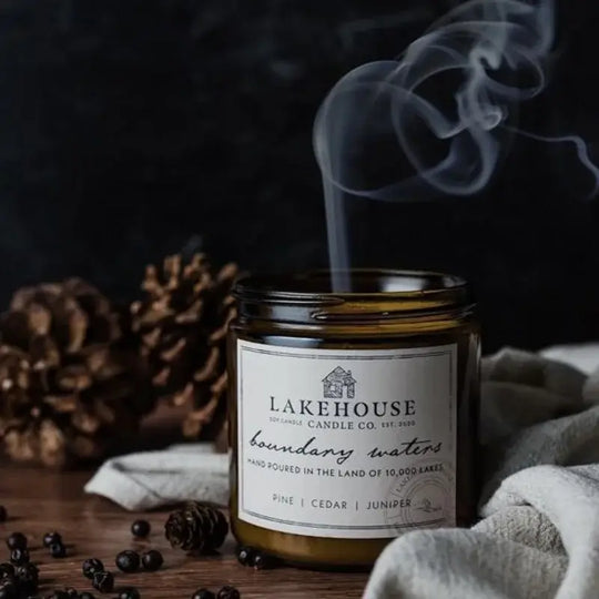 Lakehouse Candle Company Soy Candle in boundary waters with pine, cedar, juniper with a dark background with pine cones and a blanket